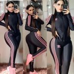 Mujer joven posando frente a un espejo en tres posiciones diferentes. Lleva un conjunto deportivo negro con tres líneas rosas en los costados de los pantalones y las mangas de la chaqueta. El conjunto tiene el logotipo de "adidas" pero está mal escrito como "adibbo" en ambas prendas. Ella usa auriculares grandes de color rosa y zapatillas deportivas rosas. Su cabello es largo y lacio, y tiene maquillaje en el rostro. Está en un espacio interior, posiblemente una habitación, con alfombra y cuadros decorativos en la pared.