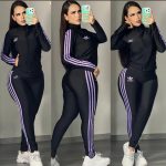 Conjunto deportivo negro con franjas lilas, modelado por una mujer en tres poses diferentes. El conjunto incluye una chaqueta y un pantalón ajustado, ambos con tres franjas lilas a lo largo de los brazos y las piernas. El logo en la chaqueta y el pantalón parece imitar el de la marca Adidas, pero el texto está escrito como "adibbo". Ella usa zapatillas deportivas blancas voluminosas y sostiene un teléfono móvil con funda verde claro mientras se toma fotos frente a un espejo. El fondo es una pared blanca y piso gris claro.