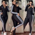 Tres fotografías de una mujer joven, posando frente a un espejo en un ambiente interior. Ella luce un conjunto deportivo negro con franjas blancas a los lados, similar a los modelos clásicos de la marca Adidas. La chaqueta tiene cremallera y las tres líneas blancas en las mangas, también se observa el logotipo en la parte superior izquierda del pecho y en la pierna superior del pantalón. Lleva zapatillas deportivas blancas y su largo cabello oscuro está peinado hacia atrás. El fondo muestra paredes claras, cuadros decorativos y una silla o puf peludo en color rosa claro. La alfombra es mullida y también de tono rosado. En cada imagen, la mujer sostiene su teléfono y se toma selfies mostrando distintas poses.